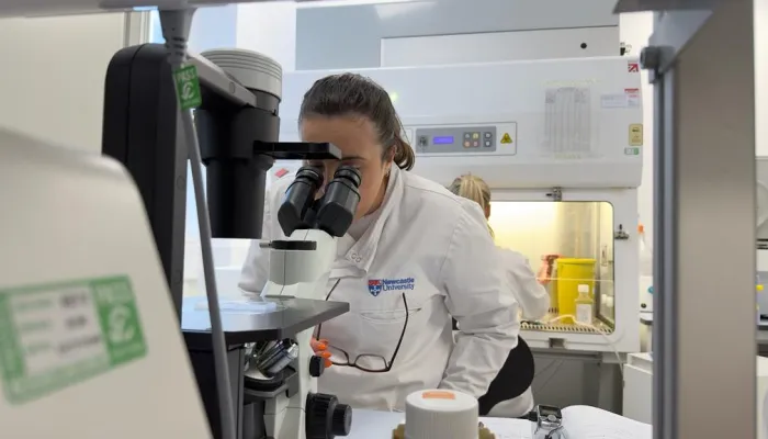 A woman in a research lab, in a white lab coat, looking into a large microscope.