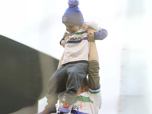 A young boy in a white and blue football t-shirt and a bright blue hat is being lifted into the air by a man in a matching shirt.