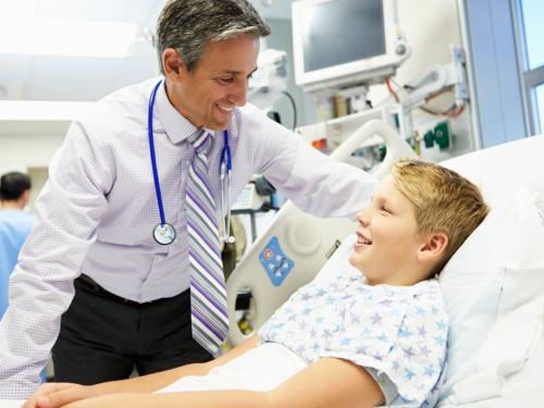 A smiling young man is lying in a hospital bed, talking to his doctor.