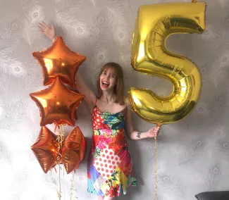 A young girl wearing a bright coloured dress is holding a number '5' gold balloon an d four orange heart balloons.