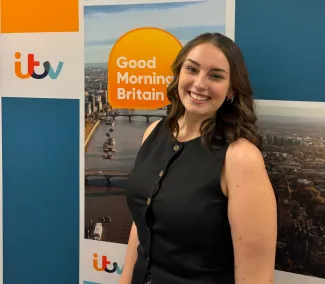 A young woman in her early 20s with brown hair, smiling and wearing a short-sleeve black shirt, stands in front of an 'ITV Good Morning Britain' backdrop.