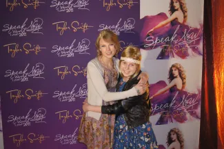 A young girl with blonde hair and a big smile is hugging Taylor Swift