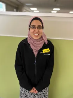 A woman wearing a pink hijab, glasses and a black fleece jacket with a yellow name badge that reads 'Hafsa' stands in front of a light green wall. She is smiling and looking directly at the camera.