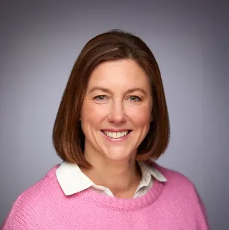 A headshot of a woman with shoulder-length brown hair. She is smiling and wearing a bright pink jumper.
