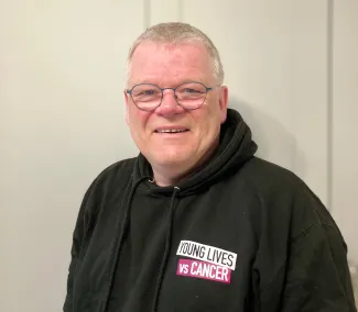A man is standing against a white background, wearing glasses and a black hoodie with the logo of Young Lives vs Cancer.