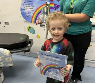 A young boy with short curly blond hair holds a ‘Congratulations’ certificate while standing beside the ‘End of Treatment Bell.’