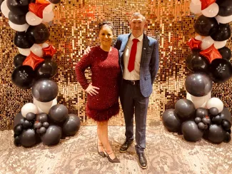 A woman and a man stand together wearing red party outfits in front of a glittery wall with balloons.