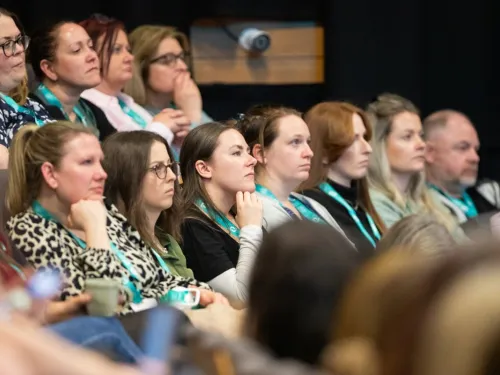 A range of people sitting in a theatre-style seating arrangement, intently listening to an out-of-shot speaker.